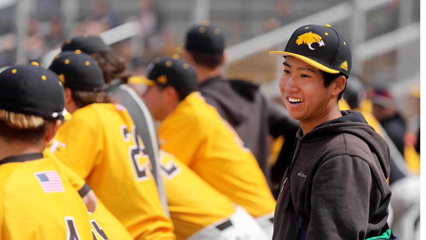 Laughing male in Taft College baseball cap with cougar emblem and black jacket looks to his left as he stands behind seven uniformed team mates in yellow shirts seen from behind and leaning on the low fence around the baseball field.