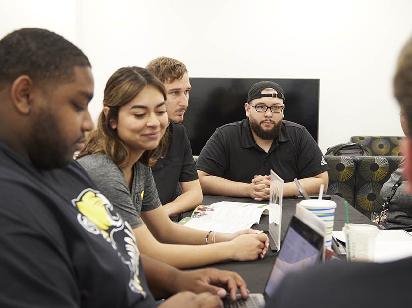 Five male and female students sit at a conference table listening to someone who is seen from the back of their head.