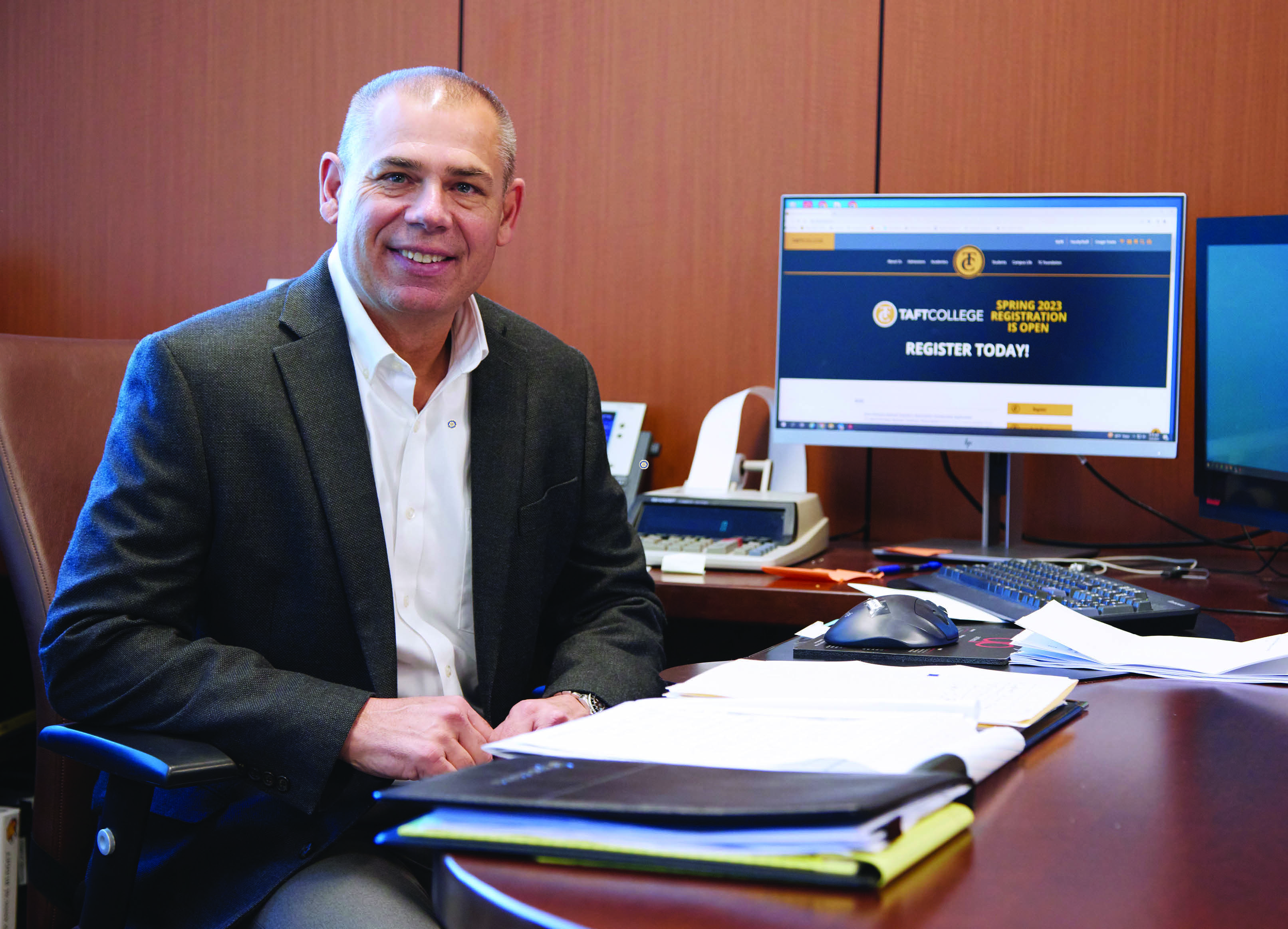 Smiling male executive in dress jacket sits at large polished computer desk in paneled office.