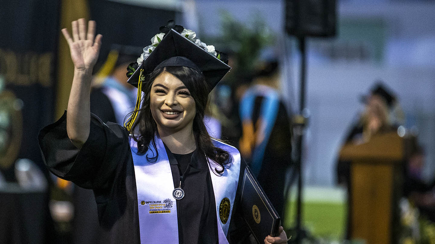 Grinning female outside in graduation cap and gown waves with her right hand and holds her certificate booklet in her left arm.