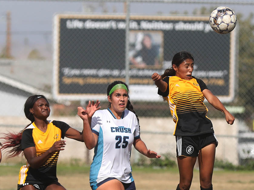 Female Taft College soccer player jumps and squints her eyes as she heads the ball away as a teammate and opponent run and watch it.