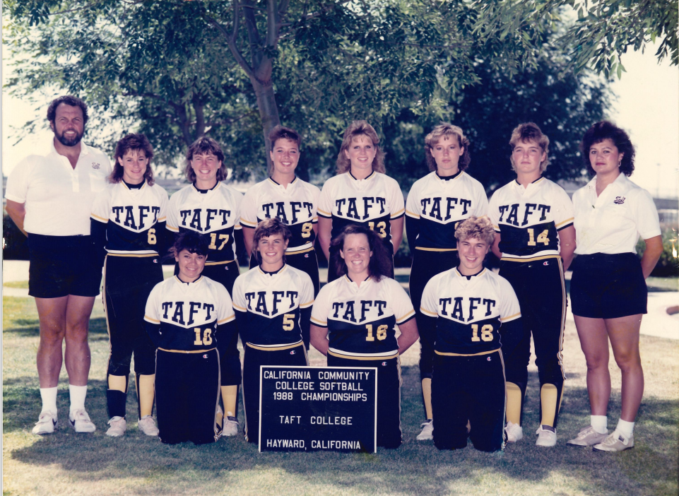 The Bandy's standing on either site of the 1998 Softball Team. They are outside, standing in 2 rows with a large green tree in the background. They are propping up a sign that says California Community College Softball Championships, Taft College Hayward, California.