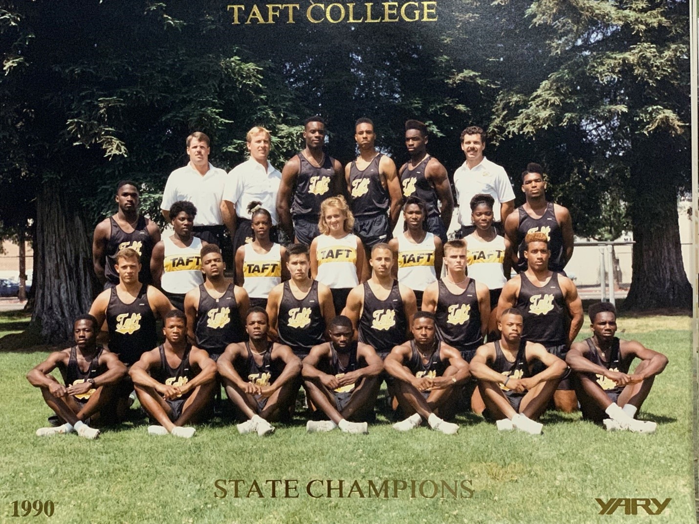 The 1990 State Championship Track Team in 3 rows. Sitting on green grass with large green trees in the background.