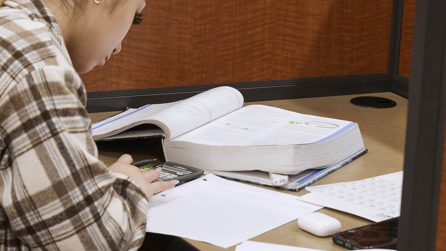 Female student with open textbook and papers across her tabletop uses a calculator.
