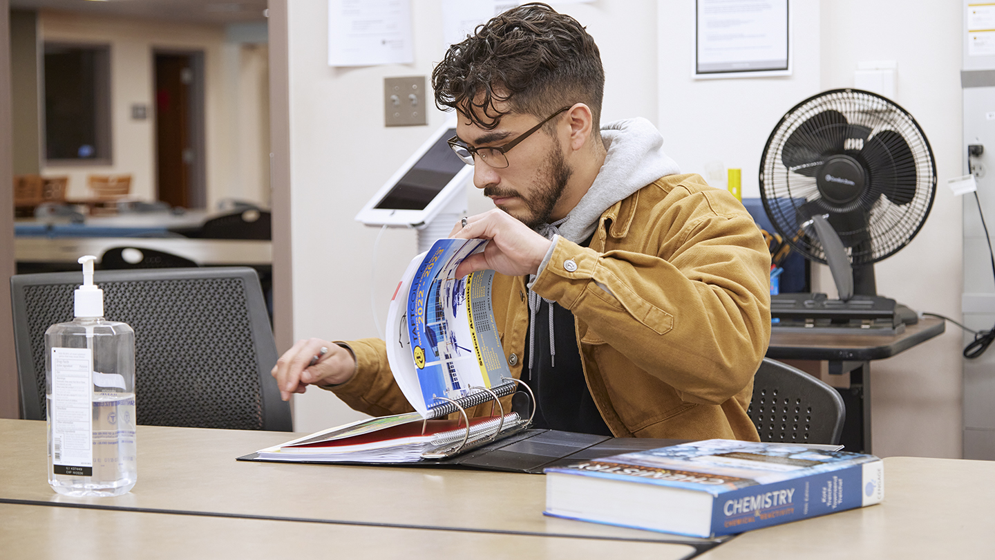 Male student in eyeglasses flips through a large ring binder at a table with a chemistry textbook.