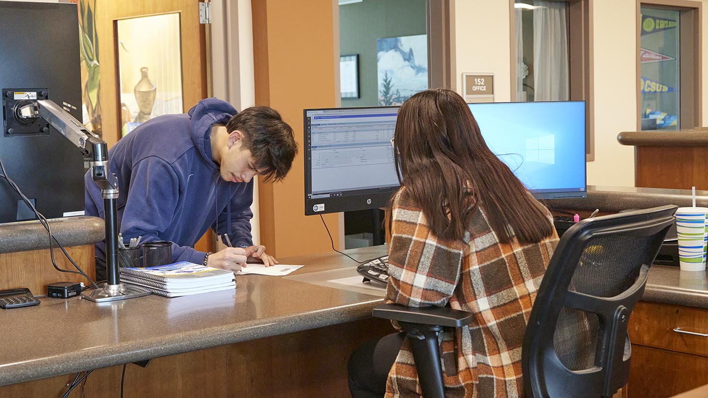 Male student leans forward and writes on a paper at a reception desk with a female seated behind it.