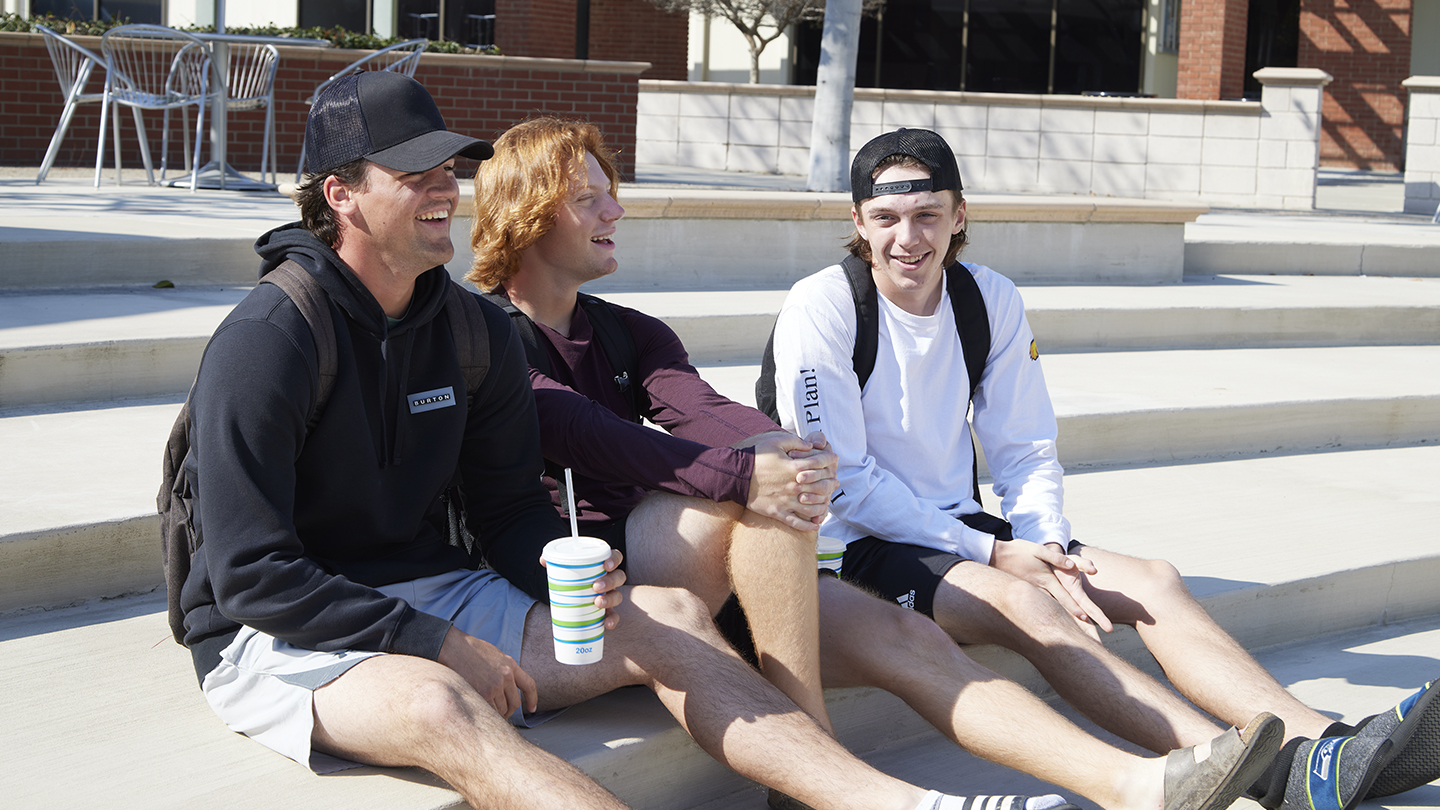 Three male students laugh while sitting on steps in the sun.
