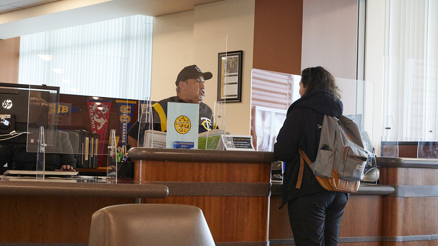 Mature male behind counter speaks with female student standing on other side of the counter.