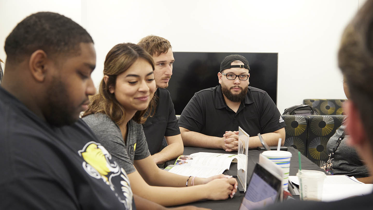 Five male and female students sit at a conference table listening to someone who is seen from the back of their head.