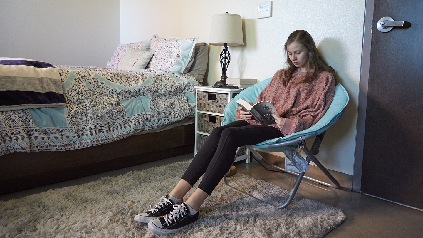 Female student reads a book in her turquoise chair, next to bed with printed bedcover and soft rug.