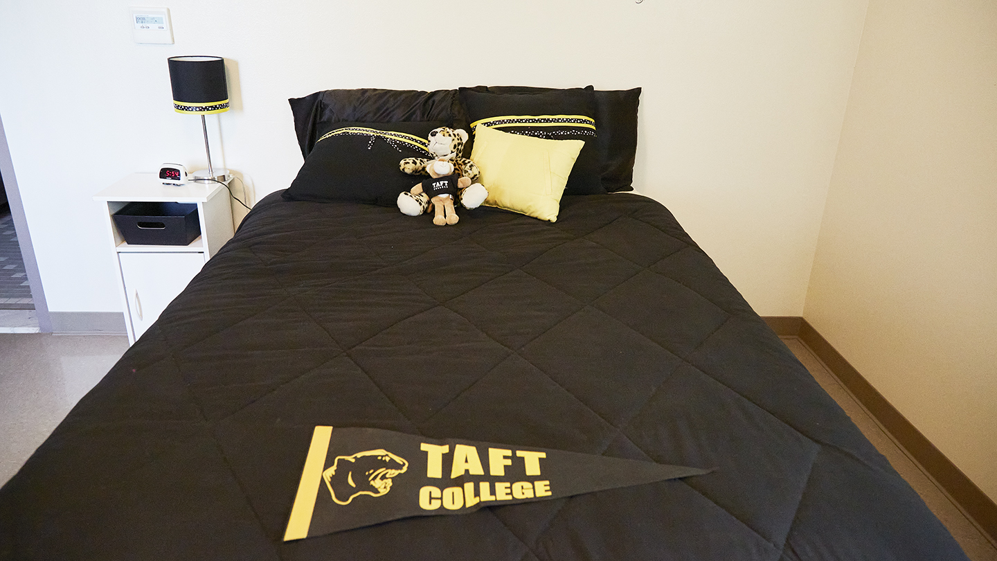 Close-up of a student bed with a black comforter, black and gold pillows, and a Taft College triangular pennant tossed on top.