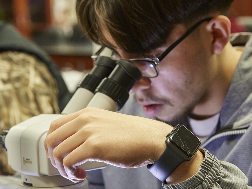 Male student with eyeglasses looks intently through the eyepiece of his microscope.
