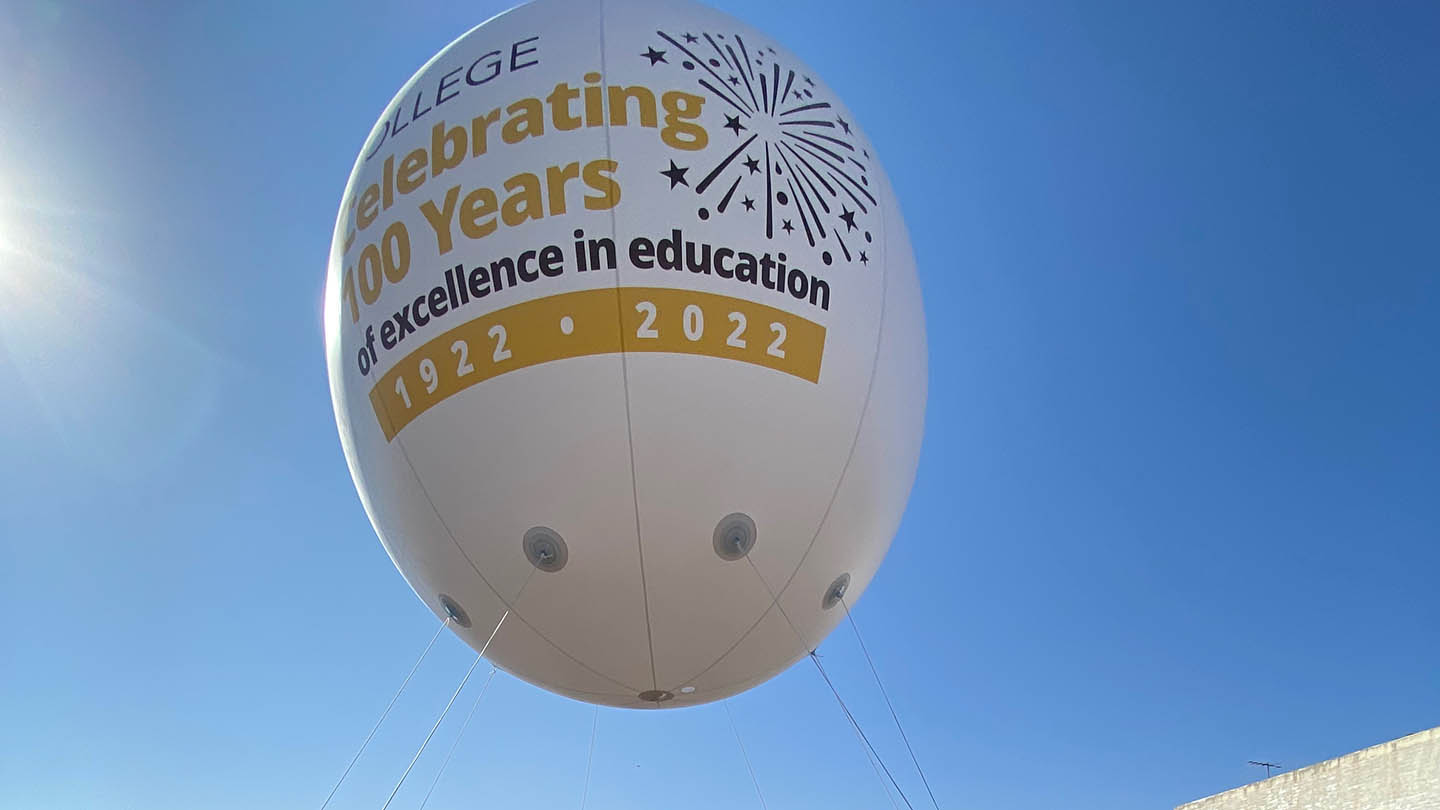 A large floating egg-shaped balloon with words about Taft College Celebrating 100 years is up in the air and guided with ropes by a team of students in yellow t-shirts.