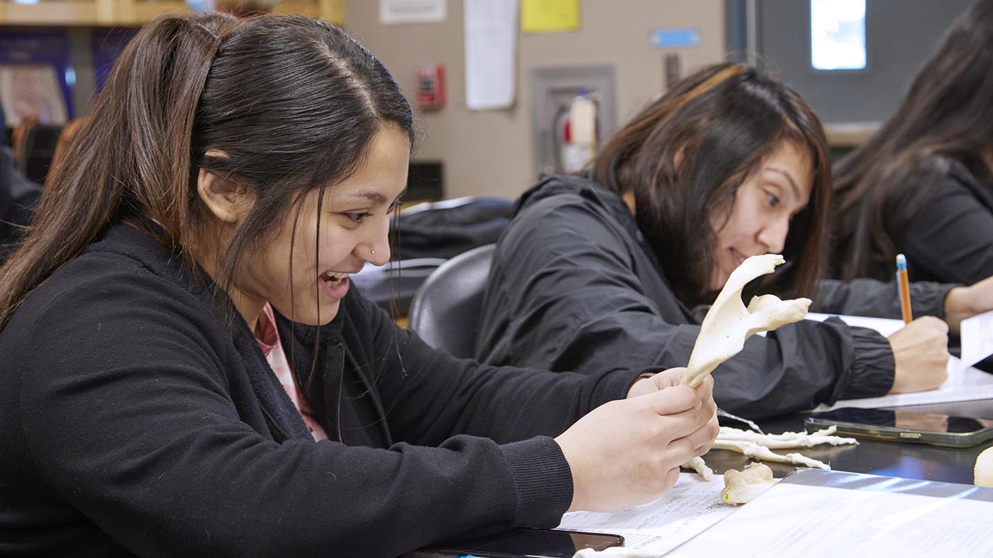 Two female students sit at a lab table. The closer one talks with a grin as she reviews her papers, while the other student focuses downward as she writes.