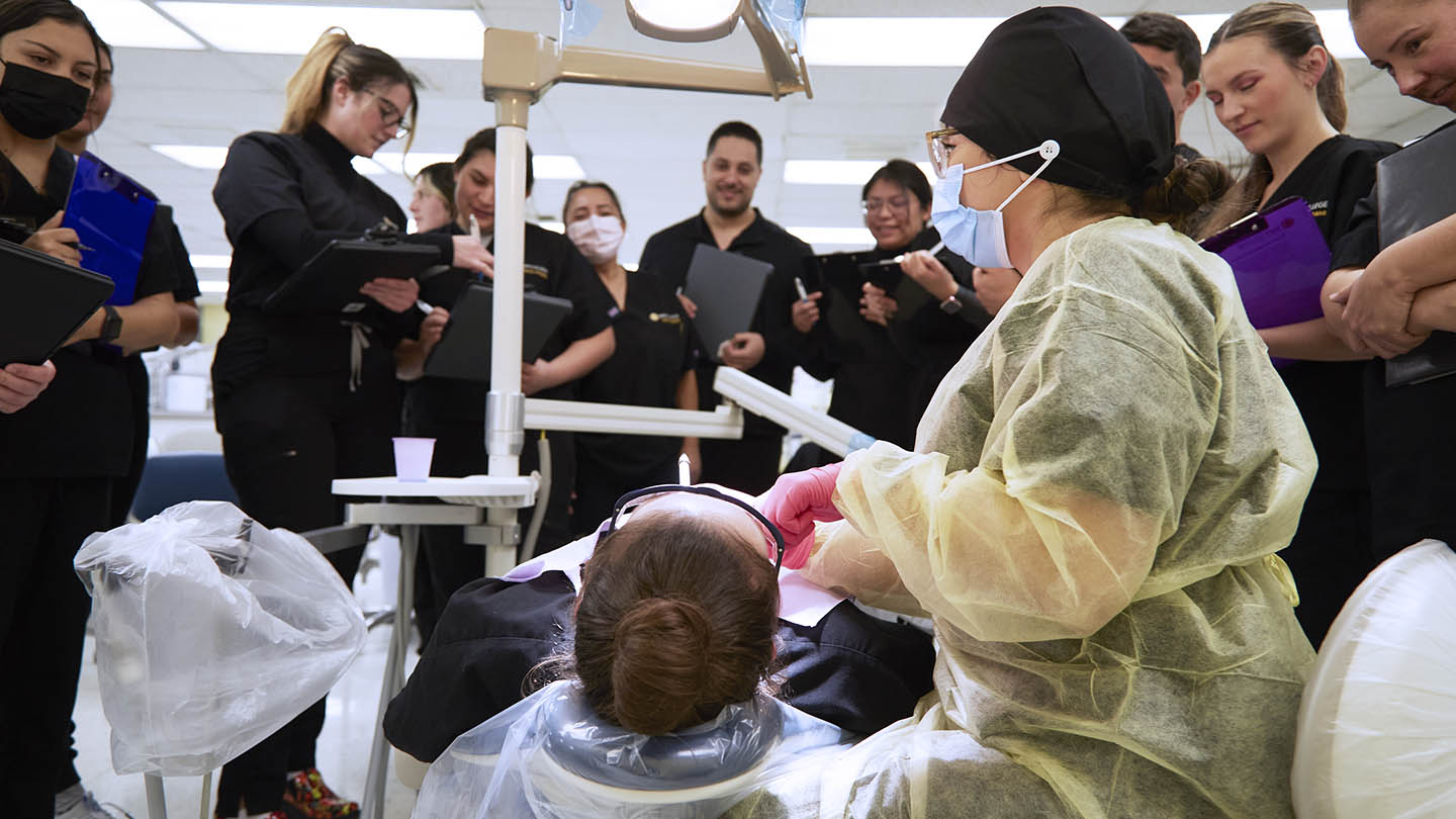 Ten students in dental scrubs form a standing semi-circle and watch closely as a seated instructor has her hands at the mouth of a person reclined in a dental chair.