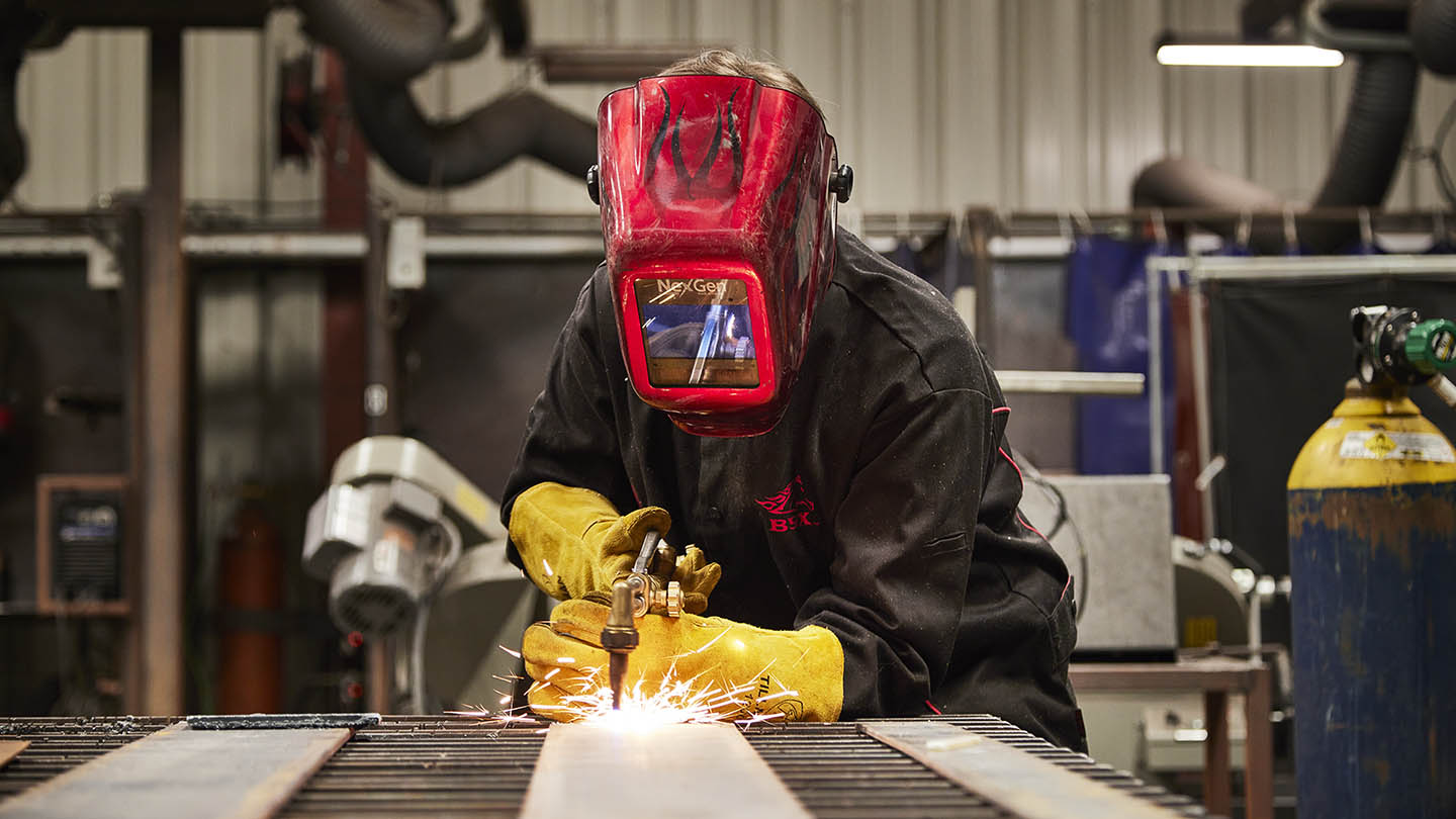 Student fully covered with dark clothing, red helmet with face shield, and yellow work gloves leans over the worktable and creates sparks with welding equipment.