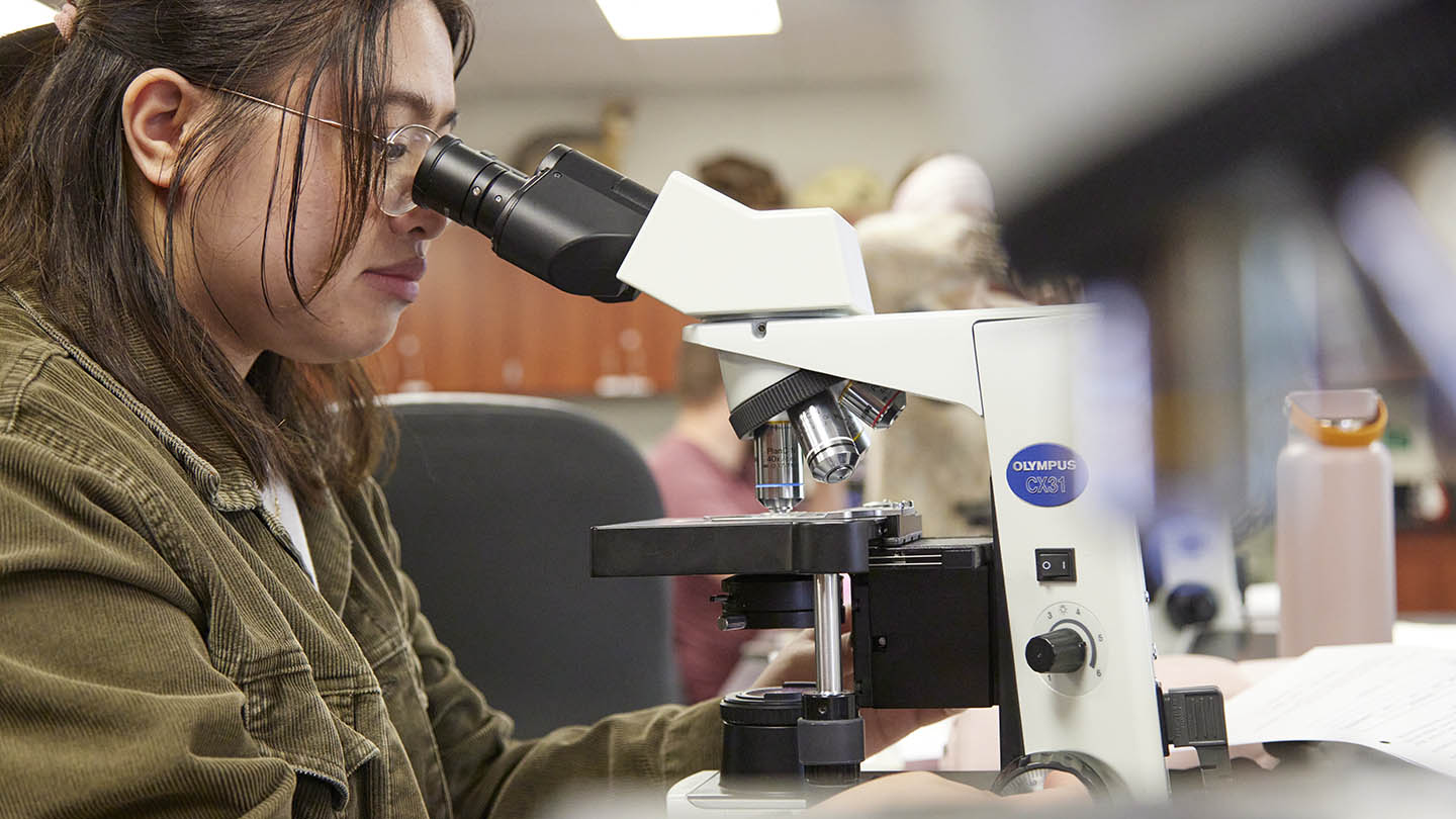 A female student with eyeglasses in a tan denim jacket looks at her specimen through a microscope.