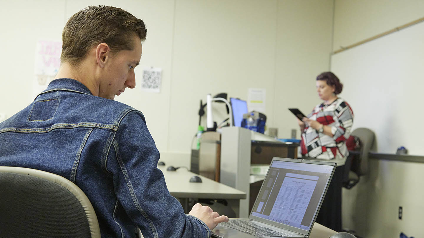 Female professor in background reviews her notes as male student in front row looks at notes on his laptop screen.