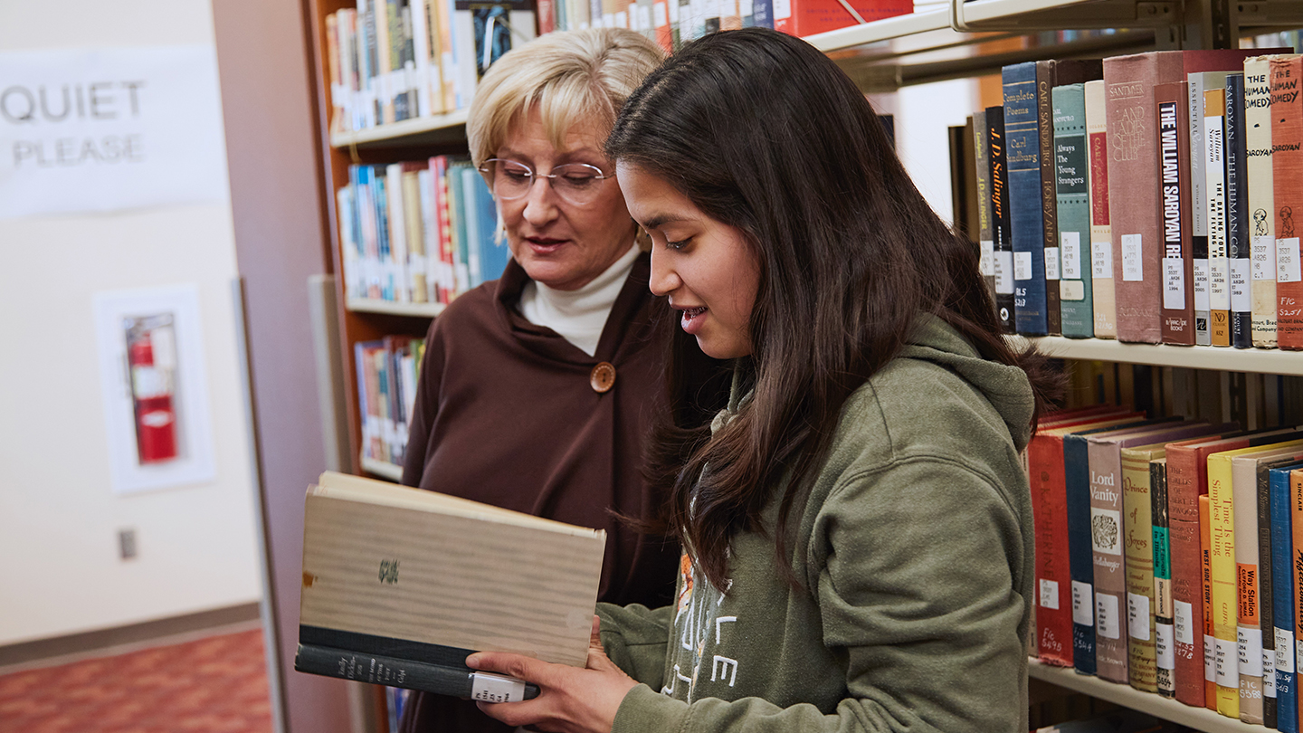Mature woman stands with female student holding a book in front of bookshelf.