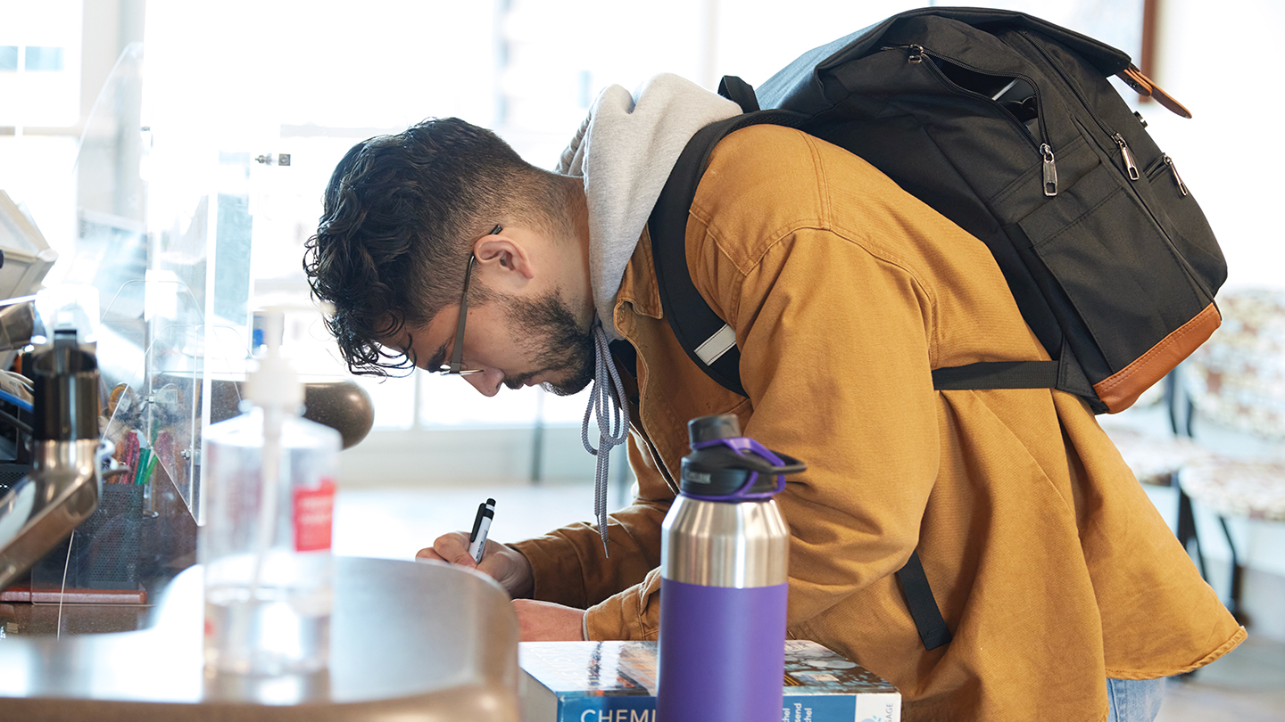 Male student in tan jacket with a chemistry textbook writes on a paper at a counter.