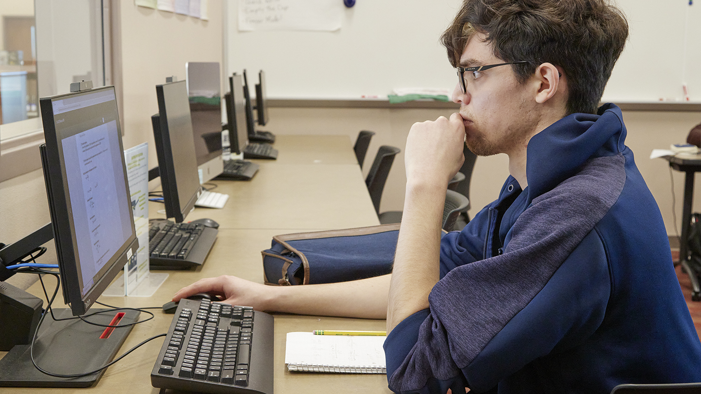 Male student with eyeglasses pensively stares at computer screen at a table with many computers.