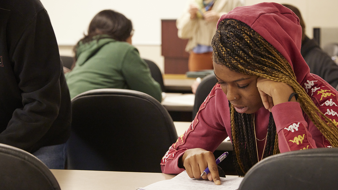 Female student in red hooded shirt sits at a table reading her notes with one elbow bent to rest her head on that hand.