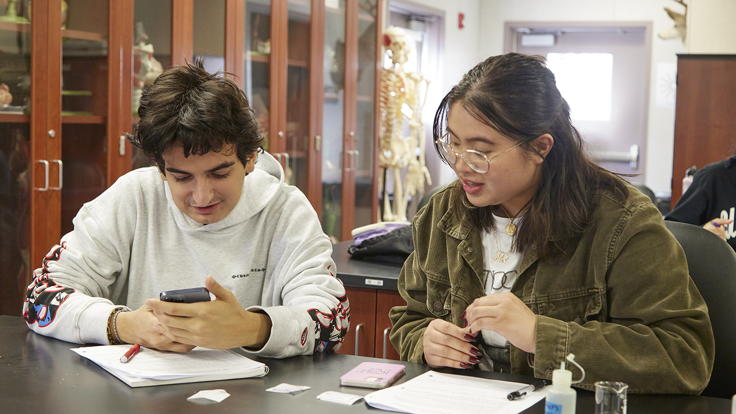 A male student and a female student, sitting at lab table, peer at hand-held equipment with a little beaker and squeeze bottle on the table.