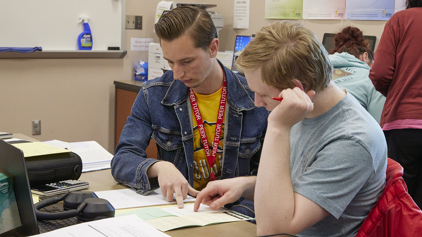 Male student with red lanyard around his neck with the words, PEER TUTOR, points to information on a paper as another male student seated next to him also points to the same page.