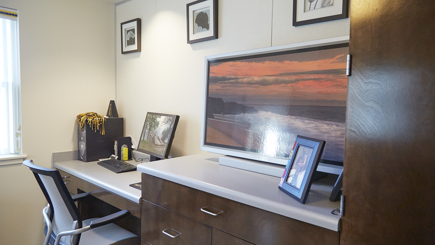 Modern styling of desk butting up to a wall with a window is adjacent to chest of drawers with a TV on top .