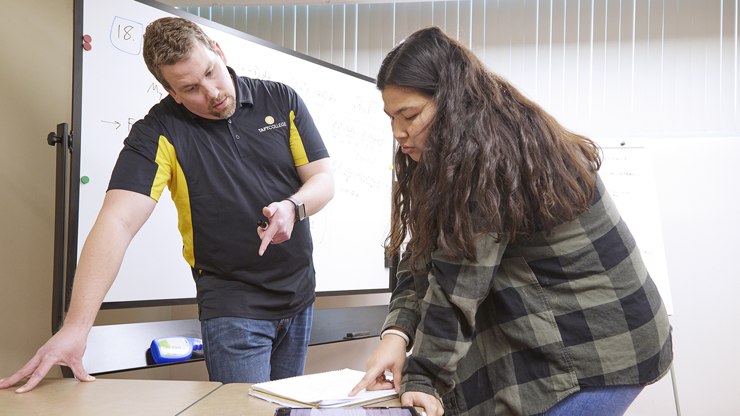 A professor gestures towards a folder on a table in the Calc Lab, as a student points towards the same folder.