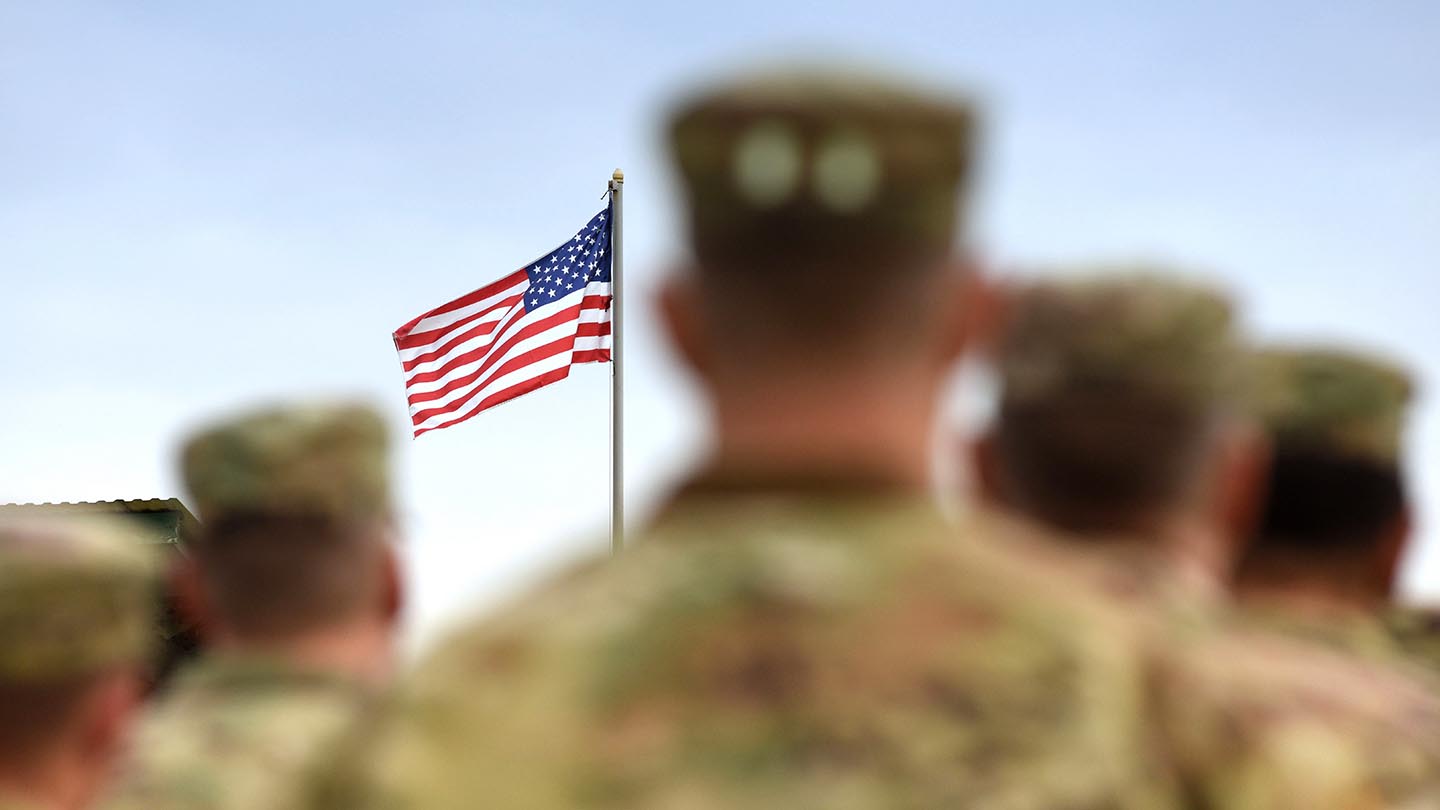Soft focus rear-view of head and shoulders of five people in camouflage military uniforms and caps in formation with a U.S. flag on pole in distance in sharp focus.